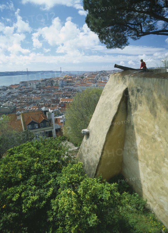 PORTUGAL, Lisbon, Visitor sitting on fortified walls beside cannon of Castelo de Sao Jorge  with rooftop view across city.
