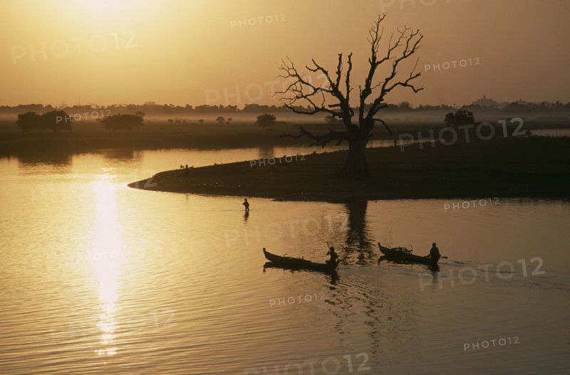 MYANMAR, Taungthaman Lake, View from U Bein Bridge at Amarapura near Mandalay with canoes silhouetted against water reflecting golden sunset and trees and landscape in drifts of mist.