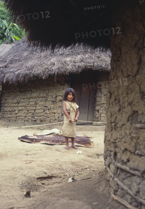 COLOMBIA, Santa Marta, Sierra Nevada, "Young Cogi girl holding cotton yarn, standing in-between two huts."
