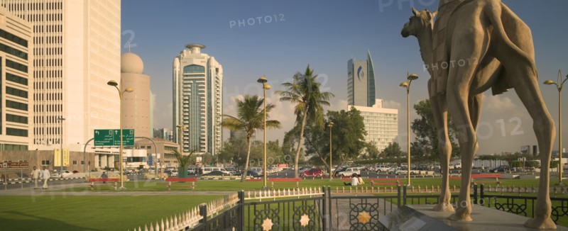 UAE, Dubai, "Camel statue in Al-Ittihad Square, people sat on grass. Palm trees and car park in distance."