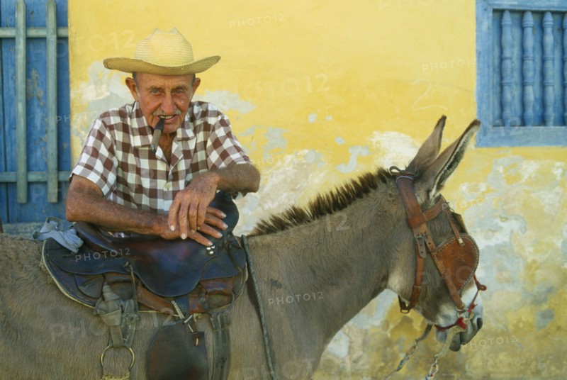 CUBA, Trinidad, Portrait of elderly Cuban man smoking a cigar and leaning on saddle of blinkered donkey.