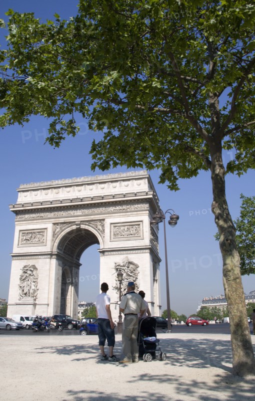 FRANCE, Ile de France, Paris, Tourist family standing in the shade of a tree in Place de Charles de Gaulle looking at the Arc de Triomphe with traffic passing around it