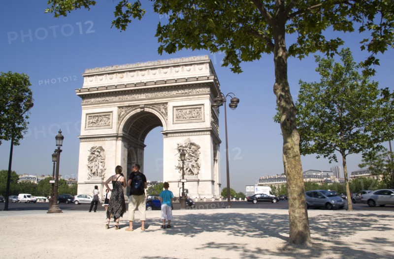 FRANCE, Ile de France, Paris, Tourist family standing in the shade of a tree in Place de Charles de Gaulle looking at the Arc de Triomphe with traffic passing around it