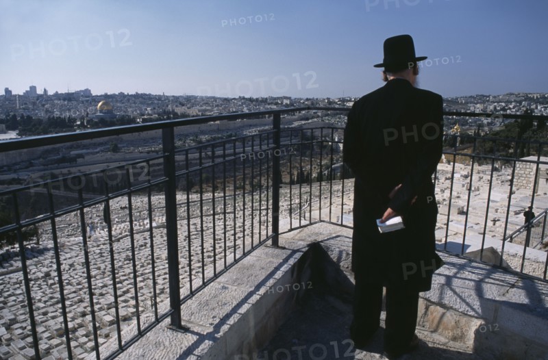 ISRAEL, Jerusalem, Elderly Ultra Orthodox Jewish man holding a bible in his hand surveying the cemetery on the Mount of Olives with the Old City in the background