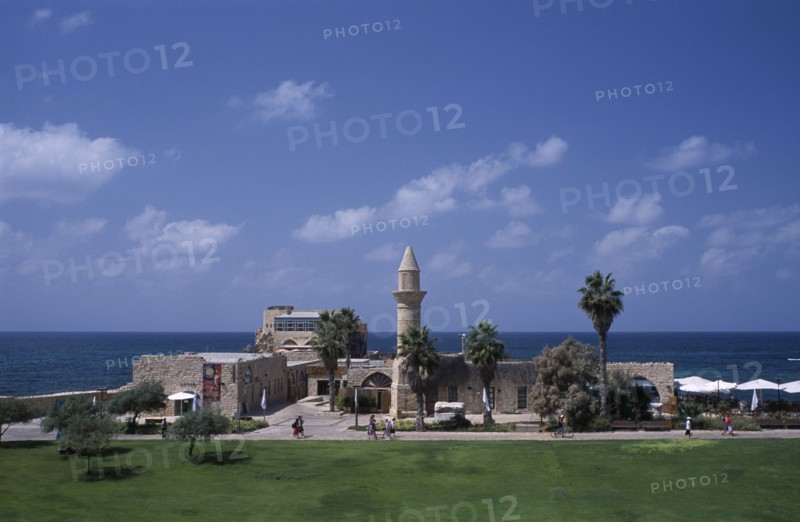 ISRAEL, Caesarea, The Herodian Harbour with eastern view of stone buildings with palm trees and visitors walking past along path.