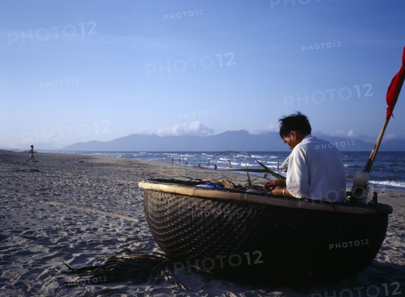 VIETNAM, Central, China Beach, Fisherman mending nets in coracle boat pulled up onto sandy beach.