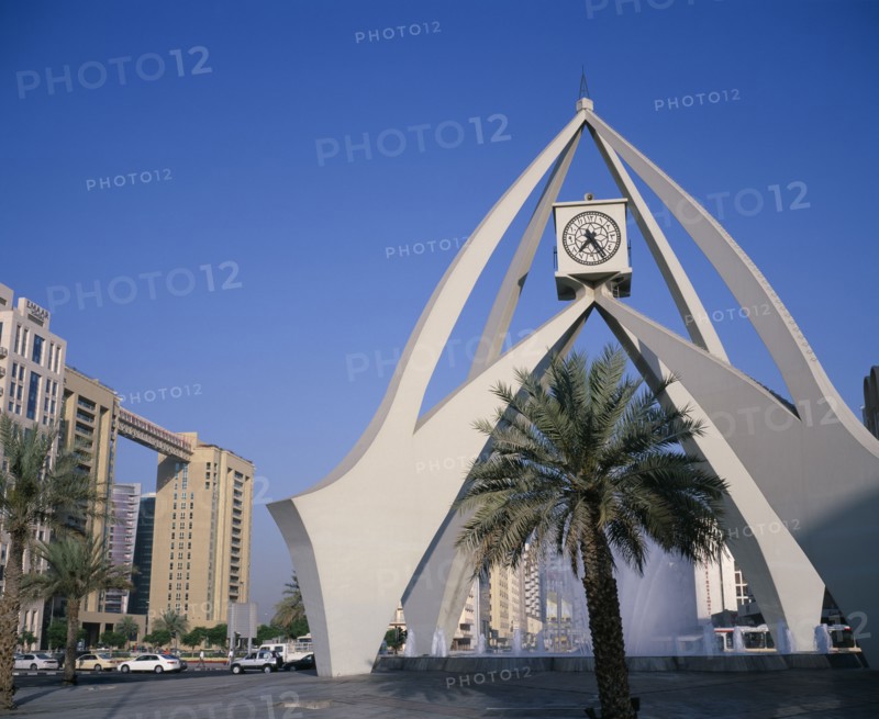 UAE, Dubai, Dubai Clock Tower.  Modern sculptural structure with pool and fountains below and palm tree in foreground.  Traffic and city buildings behind.