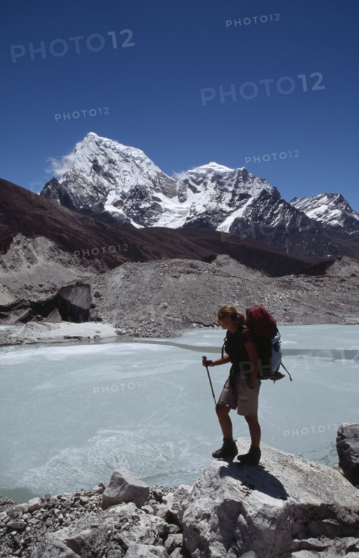 NEPAL, Himalayas, Khumbu Region, Person standing on outcrop looking over glacial lake with snow covered mountain peaks beyond on Everest Base Camp trek.