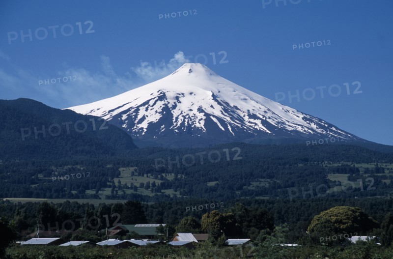 CHILE, Araucania Region, Parque Nacional Villarica , "Pucon.  Villarica Volcano 2847m.  Active volcano in the Chilean Andes, snow covered cone rising above forest covered hills with building rooftops in the foreground. "