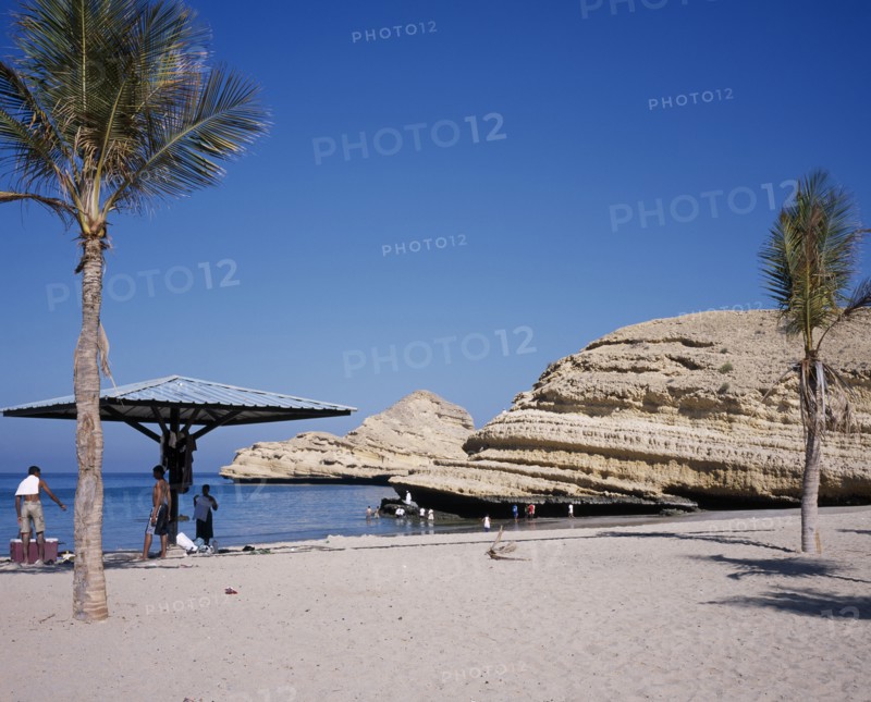 OMAN, Muscat, "Bandar Jissah beach near Al-Bustan Hotel just outside the capital city.  People at water’s edge beside eroded, rock formation with sun umbrella and palm trees."