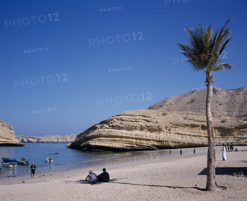 OMAN, Muscat, "Bandar Jissah beach near Al-Bustan Hotel just outside the capital city.  People at water’s edge beside eroded, rock formation with palm in foreground."