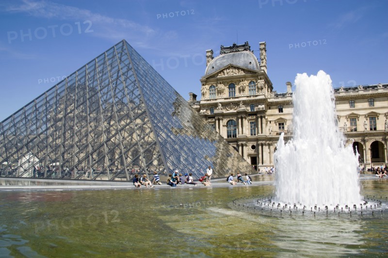 FRANCE, Ile de France, Paris, Tourists sitting beside the fountain pools in the Cour Napoleon at the Musee du Louvre with the Pyramid Entrance designed by I M Pei and the Richelieu Pavilion beyond
