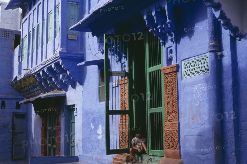 INDIA, Rajasthan, Jodhpur, Detail of blue painted house with child sitting in open doorway with green frame and decorated surround.