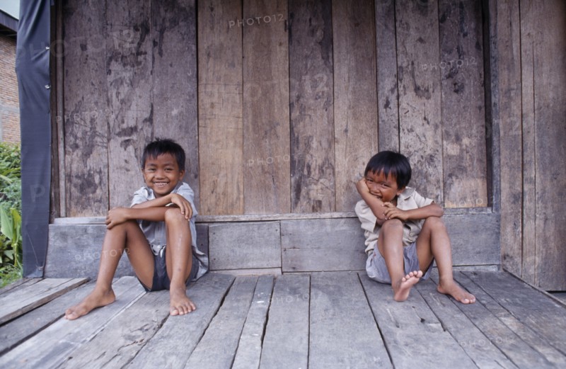 INDONESIA, Sumatra, Children, Two laughing children sitting on wooden floor of building.