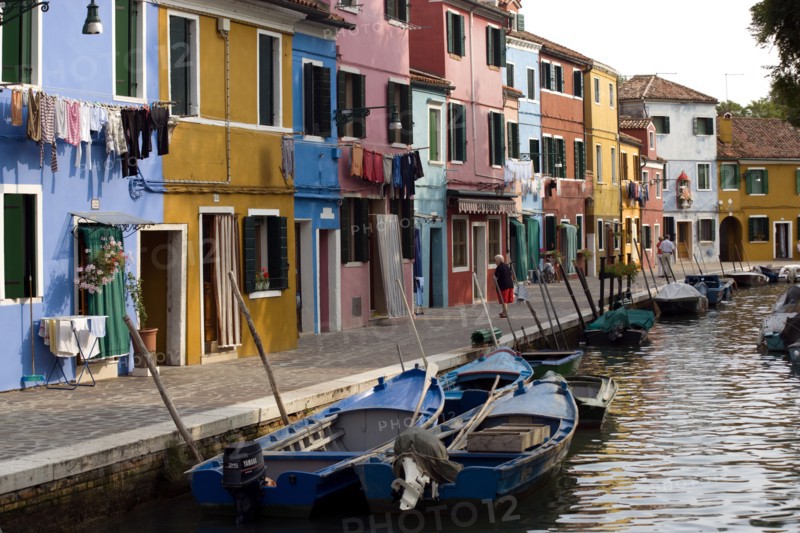 ITALY, Veneto, Venice, Colourful houses beside a canal on the lagoon island of Burano with tourists walking past boats moored alongside the edge of the canal