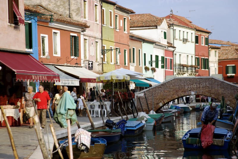 ITALY, Veneto, Venice, Colourful houses beside a canal on the lagoon island of Burano with people walking past shops and boats moored alongside the edge of the canal near to a pedestrian bridge