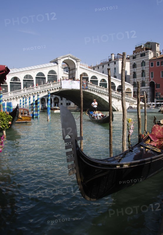 ITALY, Veneto, Venice, A gondola moored at the edge of the Grand Canal with another carrying tourists passes down river in front of The Rialto Bridge with tourist crowds