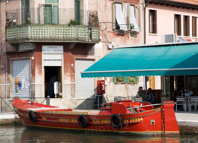 ITALY, Veneto, Venice, A traditional red lagoon barge moored alongside the Fondamenta Sebastiano Santi beside a grocery shop and coffee shop by the Ponte de la Terese bridge across the Canale di San Donato on the island of Murano