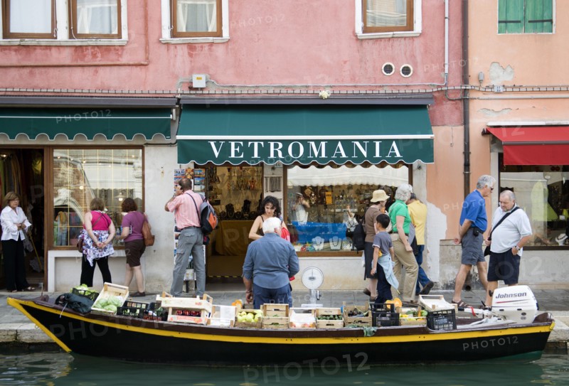 ITALY, Veneto, Venice, A fruit and vegetable vendor with his boat moored alongside the Fondamenta dei Vetrai olong the Rio dei Vetrai canal on the lagoon island of Murano. Tourists walk along the pavement looking at the shop windows