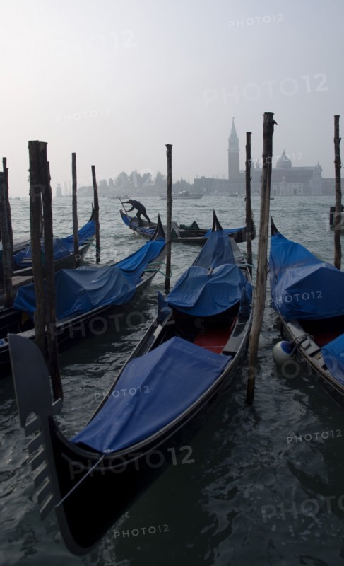 ITALY, Veneto, Venice, A gondolier works his gondola past others moored at the Molo San Marco with Palladio's church of San Giorgio Maggiore on the island of the same name in the distance on a misty day