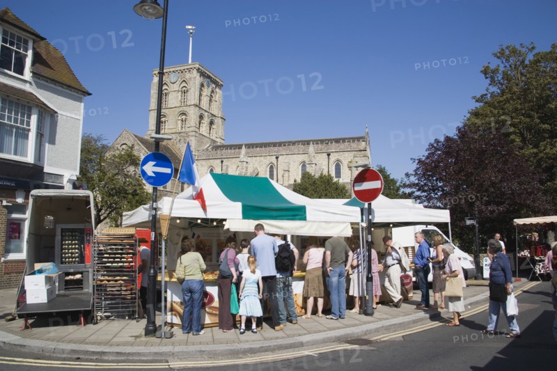 ENGLAND, West Sussex, Shoreham-by-Sea, French Market. People gathered around covered market stalls with the Church of St Mary de Haura behind.
