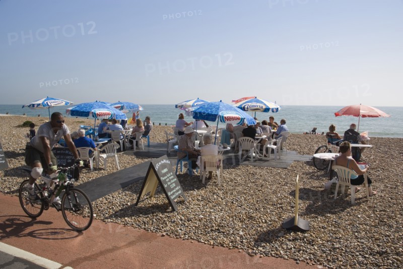 ENGLAND, West Sussex, Worthing, Beach front Cafe with people sitting at tables under parasols on the shingle overlooking the sea with a man travelling past on a bicycle