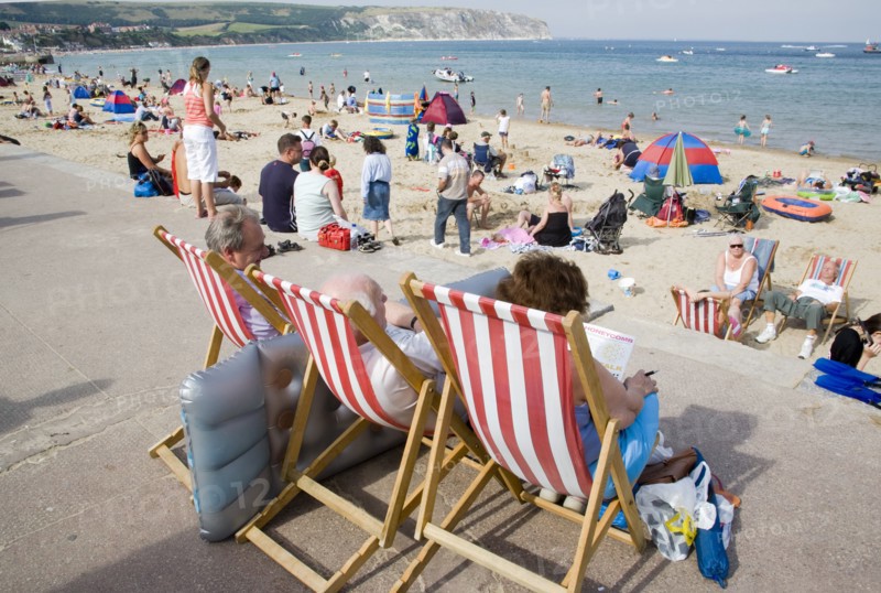 ENGLAND, Dorset, Swanage Bay, People sitting on red and white striped deckchairs looking out towards busy sandy beach with sunbathers on the sand