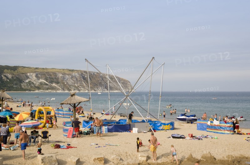 ENGLAND, Dorset, Swanage Bay, Busy colourful sandy beach scattered with inflatables and windbreaks and a bungee amusement ride next to a tiki hut in the middle