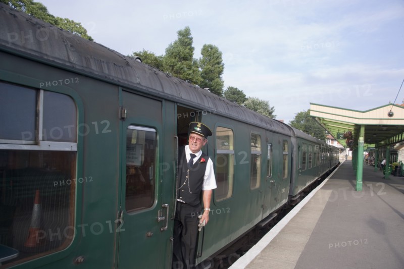 ENGLAND, Dorset, Swanage, Steam Railway Station. View along platform with train departing from station and train conductor seen looking out from carriages