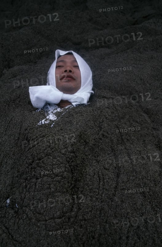 JAPAN, Kyushu, Ibusuki, Sunabulo. Hot Sand Bath with a man wearing a white towel wrapped around his head submerged in sand with only his head showing above surface