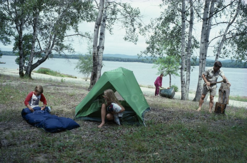 CANADA, North, Camping, "Canadian family on outback holiday setting up camp beside lake.  Children with tent and sleeping bag, mother with canoe and father using axe to split log."