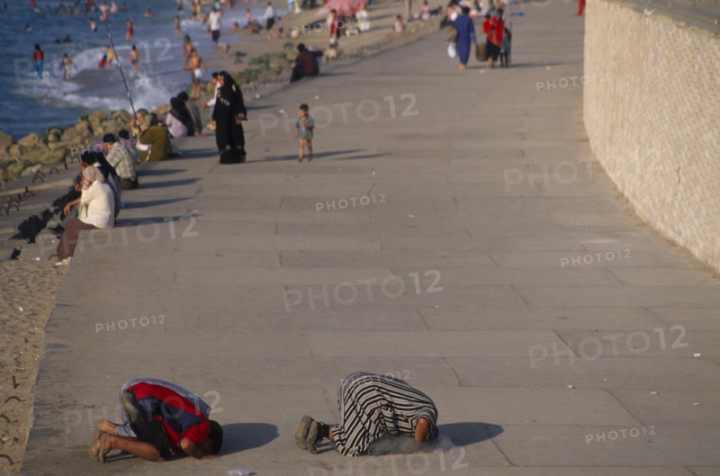 EGYPT, Nile Delta, Alexandria, Corniche Waterfront . People sitting on the edge of the promenade facing the sea with two men knelling on the ground taking part in evening prayers in the foreground.