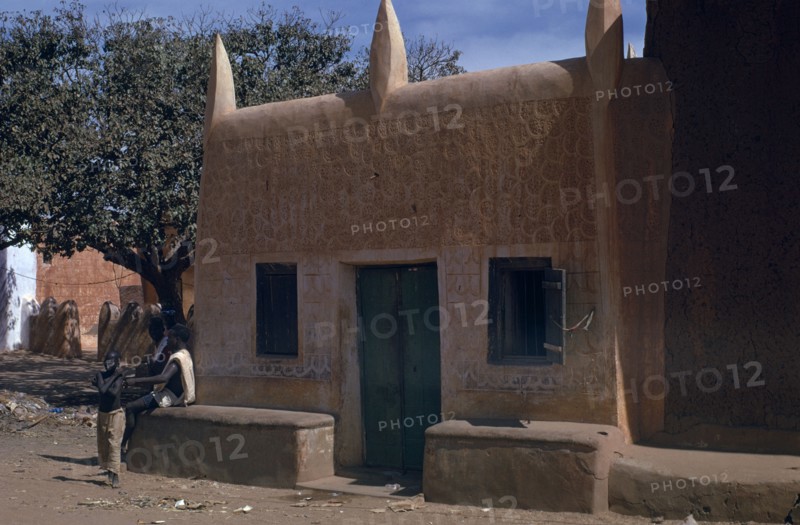 NIGERIA, Kano, Traditional mud brick Hausa dwelling with men and boy beside exterior wall.