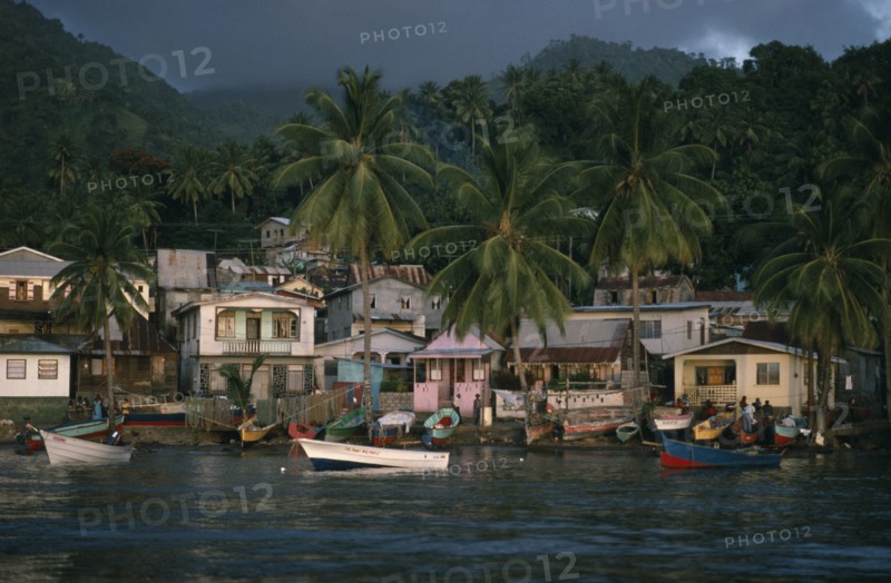 ST LUCIA, Soufriere, "Coastal housing and palm trees with colourful boats pulled up along shore.  Stormy, overcast sky above."