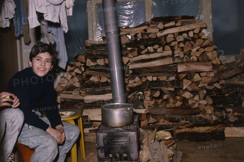 ARMENIA, Yerevan, Boy sitting beside wood burning stove in house with large stack of wood behind during winter time.