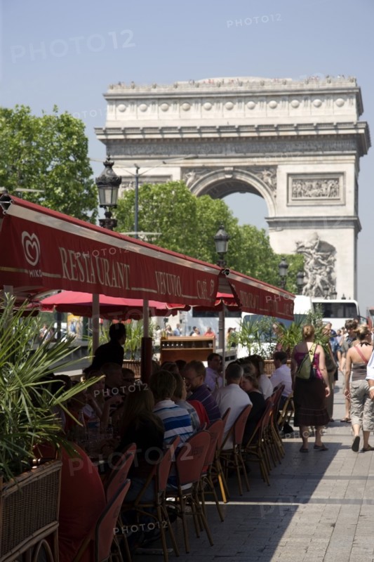 FRANCE, Ile de France, Paris, Tourists eating lunch at tables under shade in the Champs Elysees with the Arc de Triomphe in the distance