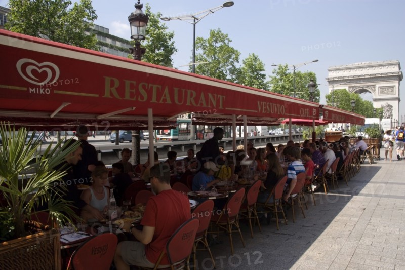 FRANCE, Ile de France, Paris, Tourists eating lunch at tables under shade in the Champs Elysees with the Arc de Triomphe in the distance