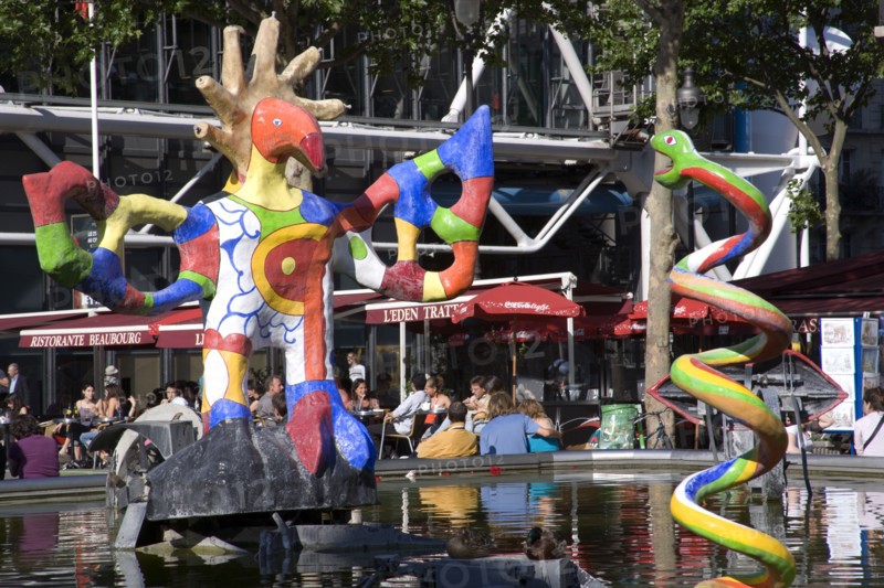 FRANCE, Ile de France, Paris, People sitting at tables under umbrellas beside the colourful contemporary fountains by Niki de Saint Phalle and Jean Tinguely in Place Igor Stravinsky beside the Pompidou Centre in Beauborg Les Halles