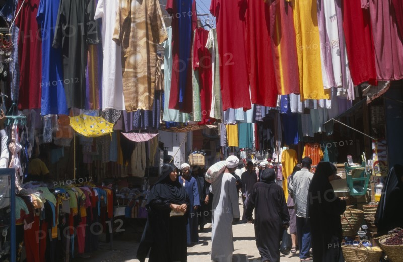 EGYPT, Nile Valley, Luxor, Sharia el-Souk street in the main market / bazaar. Men and women walking amongst market stalls under hanging displays of brightly coloured clothing