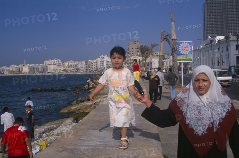 EGYPT, Nile Delta, Alexandria, Corniche Waterfront. A mother wearing an Islamic headscarf holding her daughters hand while she walks along wall above her.