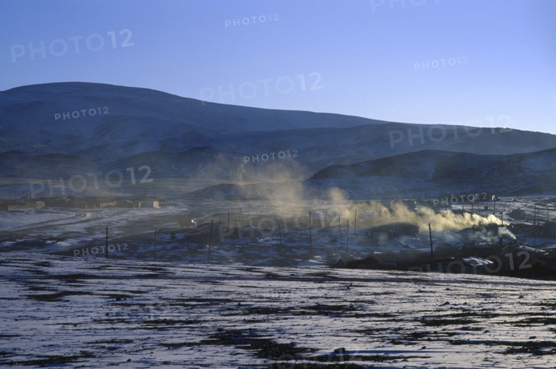 MONGOLIA, Hovd Province, Erdeneburen, Drifting smoke from buildings in landscape with light covering of snow in March sunset.