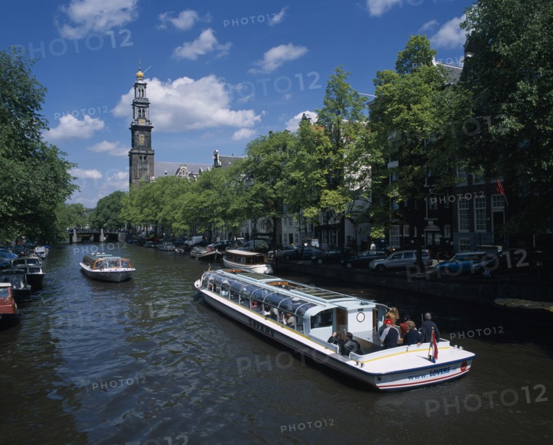 HOLLAND, Noord-Holland, Amsterdam, Pleasure boats on canal lined by trees and parked cars on side streets overlooked by typical architecture partly seen through branches.  Distant bridge and bell tower.