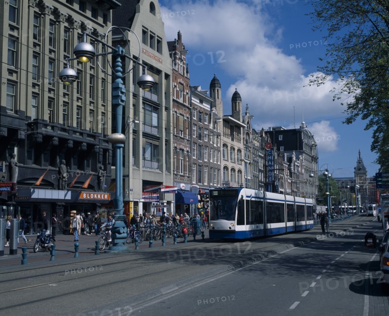 HOLLAND, Noord-Holland, Amsterdam, "Damrak.  Busy street scene, lined with shop, bar and hotel facades.  Crowds of pedestrians, parked bicycles, marked cycle lane and blue and white tram."