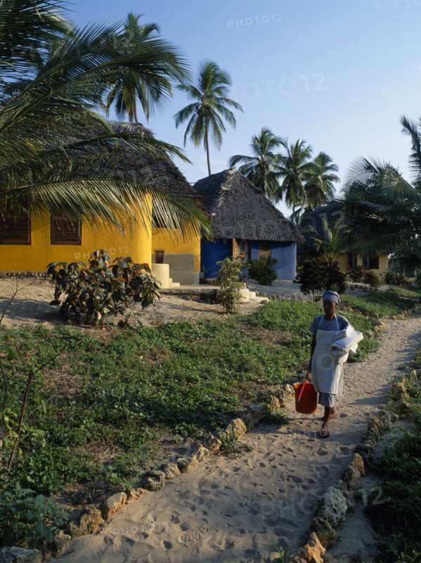 TANZANIA, Pangani, The Tides Hotel.  Brightly painted thatched huts amongst palm trees with maid carrying cleaning equipment and fresh towels on path in foreground.