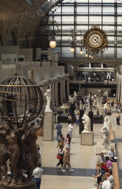 FRANCE, Ile de France, Paris, "Musee d’Orsay interior, visitors walking amongst sculpture inside large gallery space with ornate lighting and clock above."
