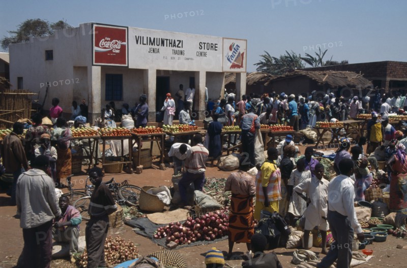 MALAWI, Jenda, "Crowded market scene, fruit and vegetable stalls with trading centre building behind."