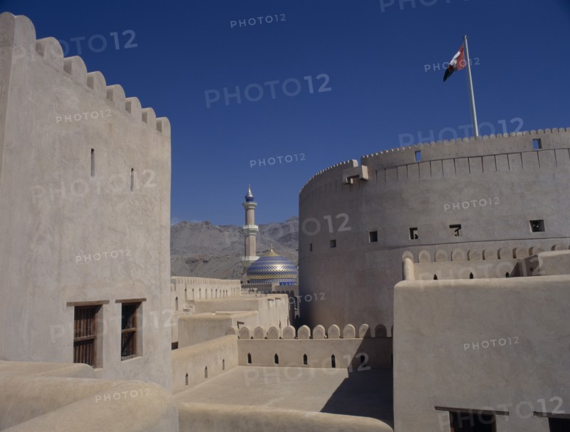 UAE, Oman, Nizwa, Minaret and blue and gold domed roof of mosque seen from fort walls with barren mountainside behind.