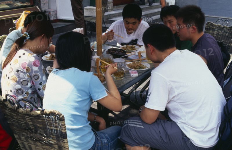 CHINA, Beijing, "Wangfujing shopping street.  Group of young people using chopsticks to eat meal, and drinking at outside table."
