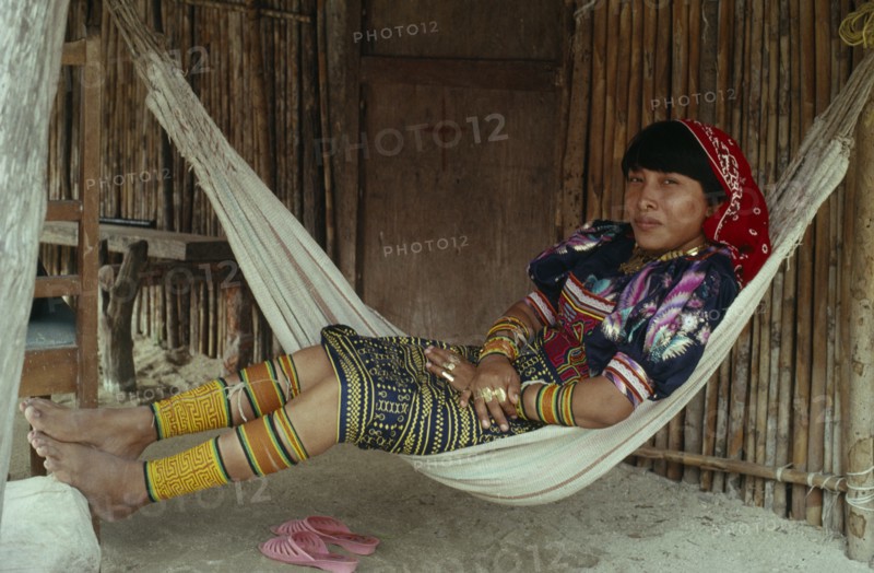 PANAMA, San Blas Islands, Kuna Indians, Young Kuna mother rests in hammock outside home  wearing brightly coloured traditional layered applique mola panel    traditional bead design arm and leg bands and gold nose ring. Cuna Caribbean American Central America Classic Classical Colored Female Women Girl Lady Hispanic Historical Latin America Latino Older Panamanian West Indies