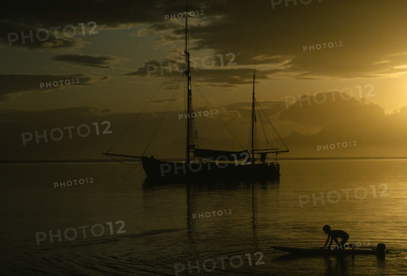 PACIFIC ISLANDS, French Polynesia, Tahiti, Sunset reflected in still expanse of water with silhouetted sail boat at anchor and children playing in the water in foreground.